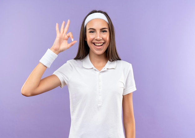 Vrolijke, vrij sportieve meid met een hoofdband en een polsbandje dat een goed teken doet dat op een paarse muur met kopieerruimte wordt geïsoleerd