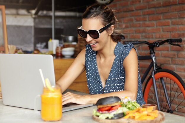 Vrolijke vrouw die in modieuze zonnebril op laptop computer intikken