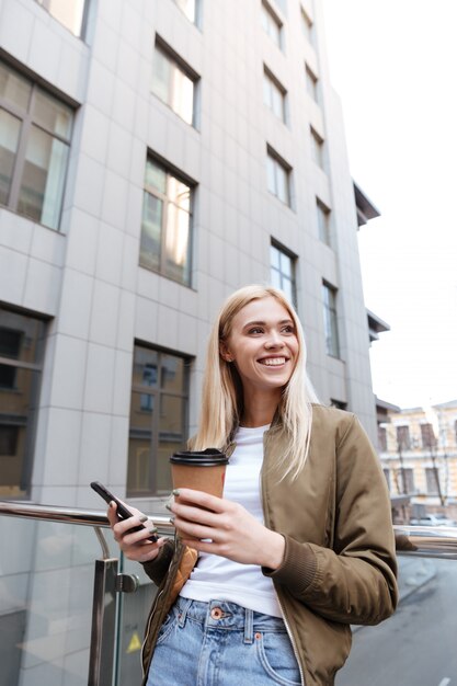 Vrolijke vrouw die met kop van koffie en smartphone opzij kijken