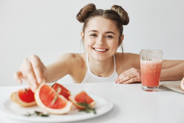 Vrolijke vrouw die nemend plak van grapefruit van plaatzitting bij lijst over witte muur glimlachen. Gezond fitness eten.