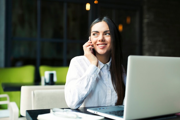 Vrolijke vrouw die op telefoon bij laptop spreekt