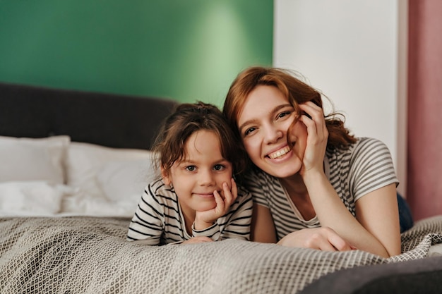 Vrolijke vrouw en haar schattige dochter liggen op bed en kijken naar de camera met een glimlach