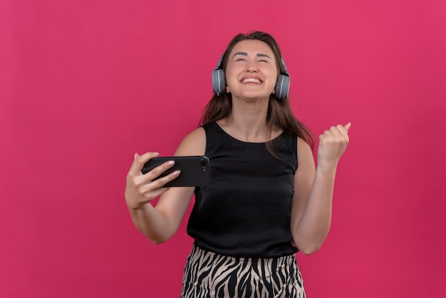 Vrolijke vrouw, gekleed in een zwart hemd, luistert muziek uit een koptelefoon op een roze muur