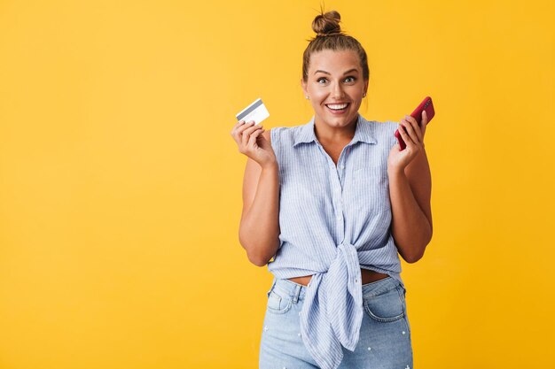 Vrolijke vrouw in shirt die vreugdevol in de camera kijkt terwijl ze creditcard en mobiel in handen houdt over gele achtergrond