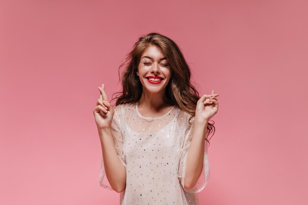 Vrolijke vrouw kruist vingers en glimlacht breed op roze achtergrond Jonge dame in witte jurk doet wens en poseert op geïsoleerde
