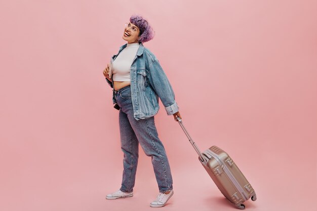 Vrolijke vrouw met bril en licht T-shirt en strakke jeansstappen en koffer houdt. Vrolijke vrouw met paars haar poseren.