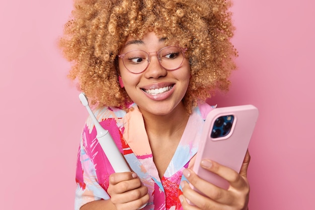 Vrolijke vrouw met krullend haar controles ontvangen bericht op smartphone terwijl tandenpoetsen houdt elektrische tandenborstel draagt bril en shirt geïsoleerd over roze achtergrond hygiëne routines