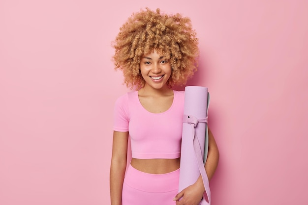 Gratis foto vrolijke vrouw met krullend haar draagt sportieve outfit houdt opgerolde mat klaar voor fitnesstraining thuis geïsoleerd op roze achtergrond gelukkig sportief vrouwelijk model draagt karemt voor yogapraktijk