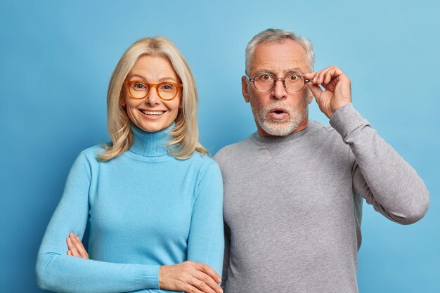 Vrolijke vrouw van middelbare leeftijd en haar gepensioneerde echtgenoot reageert op schokkend nieuws houdt de hand op bril geïsoleerd over blauwe muur
