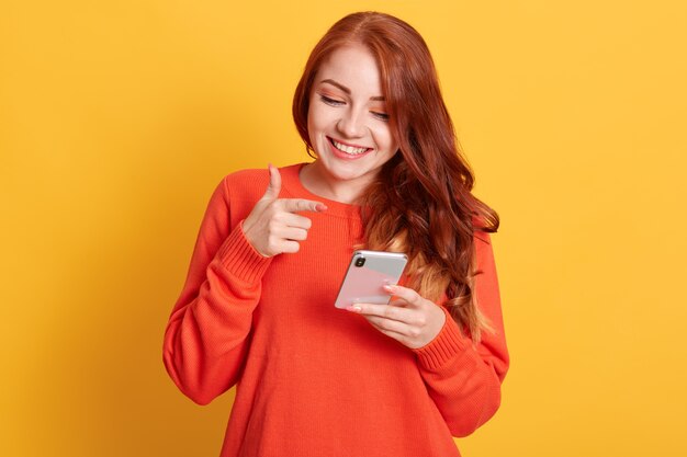 Vrolijke vrouw wijzend naar haar slimme telefoonscherm met wijsvinger, mobiele telefoon kijken met een charmante glimlach