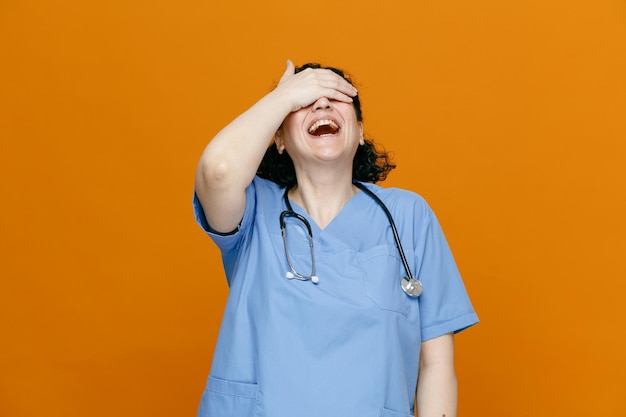 Vrolijke vrouwelijke arts van middelbare leeftijd die uniform en stethoscoop om haar nek draagt en hand op ogen houdt lachend geïsoleerd op oranje achtergrond