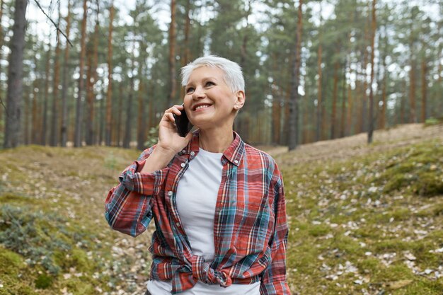 Vrolijke vrouwelijke gepensioneerde m / v met kort blond haar poseren in de wilde natuur met pijnbomen op de achtergrond, genieten van versheid, indrukken delen met vriend, spreken op mobiele telefoon, lachen