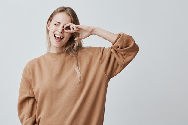 Vrolijke vrouwelijke vrouw met blond steil haar, verheugt zich met succes voor examens, blij met een ontmoeting met groepsgenoten, breed glimlachend. Opgetogen mooie blonde vrouw heeft een speelse uitstraling