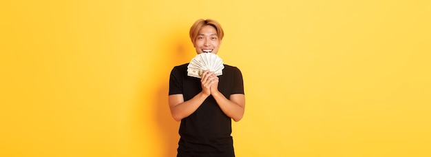 Gratis foto vrolijke winnende knappe aziatische man die tevreden glimlacht en geld vasthoudt en zich gelukkig voelt als hij geel staat