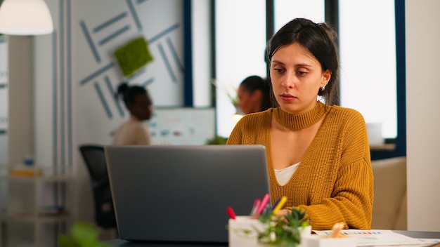 Vrolijke zakendame typen op laptopcomputer en glimlachend zittend aan een bureau in een druk opstartkantoor dat geniet van werk op een creatieve werkplek. Divers team analyseert statistische gegevens in modern bedrijf