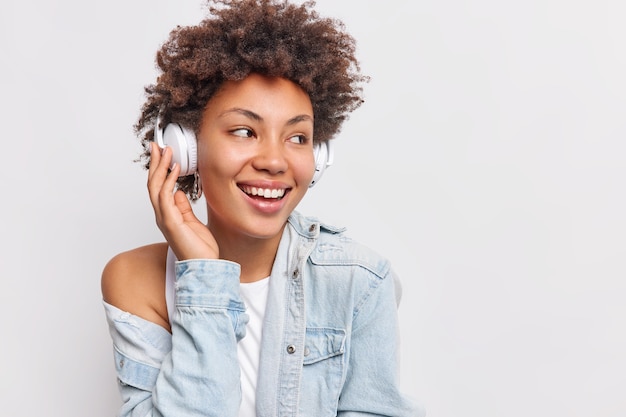 Vrolijke zorgeloze vrouw met krullend haar kijkt weg glimlacht tandjes draagt draadloze stereo hoofdtelefoon op oren luistert favoriete muziek uit afspeellijst geniet van goede geluidskwaliteit poseert over witte muur vrije ruimte