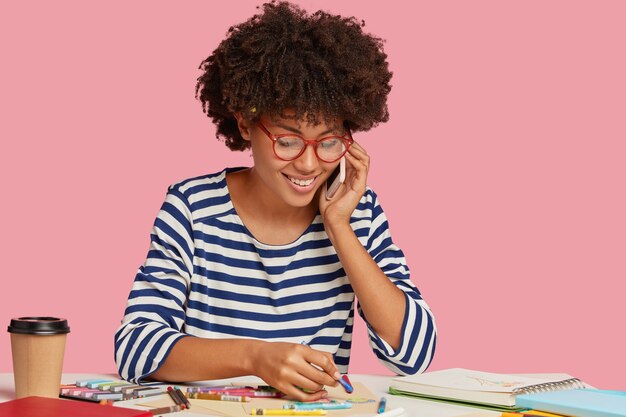 Vrolijke zwarte dame geniet van schilderen, maakt een foto op een blanco vel papier, draagt een optische bril, voert een telefoongesprek, glimlacht zachtjes als iets aangenaams bespreekt, geïsoleerd op roze muur