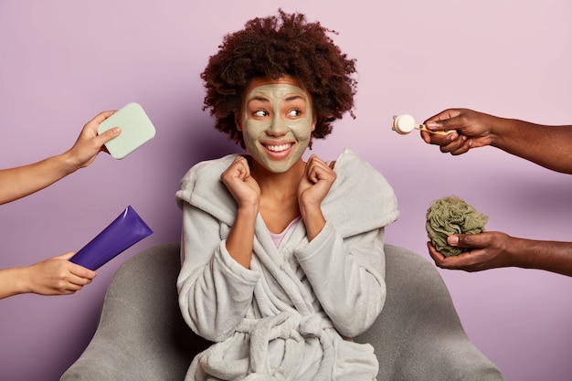 Vrolijke zwarte vrouw ontspant met gezichtsmasker van klei, ondergaat huidverzorgingsprocedure