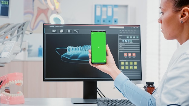 Vrouw aan het werk als tandarts met groen scherm op smartphone in mondverzorgingskantoor. Tandarts met mobiele telefoon met mockup-sjabloon en geïsoleerde achtergrond voor tandverzorging en tandheelkunde.
