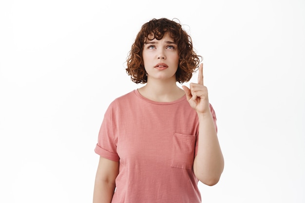 vrouw aarzelend wenkbrauwen optrekkend, wijzend en opkijkend met een twijfelachtig, ondervraagd gezicht, kopieerruimte bovenop lezend, staand in t-shirt op wit.