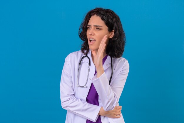 vrouw arts dragen witte jas met een stethoscoop schreeuwen iemand bellen met de hand in de buurt van de mond op geïsoleerde blauw