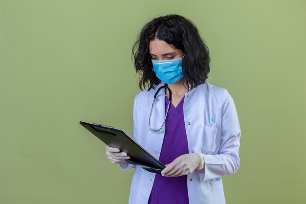 vrouw arts dragen witte jas met stethoscoop in medische beschermend masker kijken naar klembord in haar handen met ernstige geconcentreerde gezicht staande op geïsoleerde groen