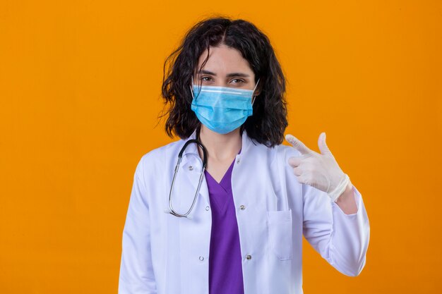 vrouw arts dragen witte jas met stethoscoop in medische beschermend masker met zelfverzekerde glimlach wijzend met vinger naar haar masker staande op geïsoleerde oranje