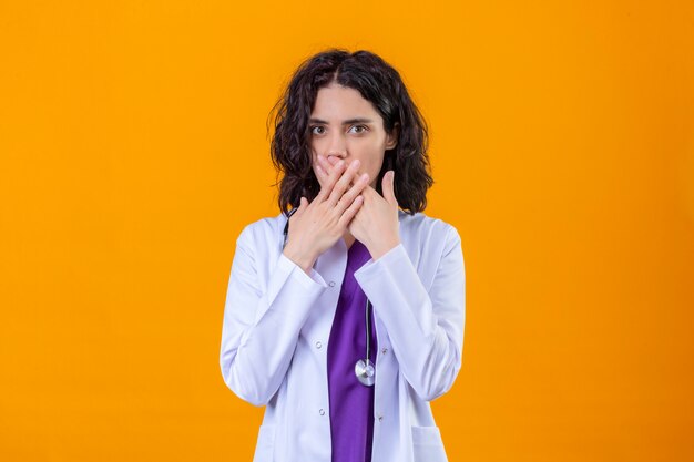 vrouw arts dragen witte jas met stethoscoop op zoek verrast kegelvormige mond met handen staande op geïsoleerde oranje