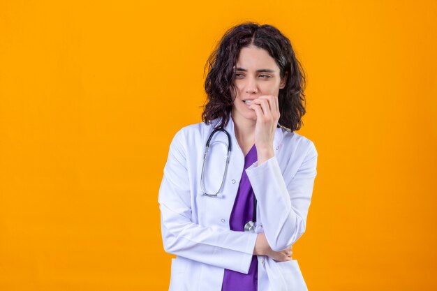 vrouw arts dragen witte jas met stethoscoop opzij kijken met verdachte uitdrukking nerveus vingernagels bijten staande op geïsoleerde oranje