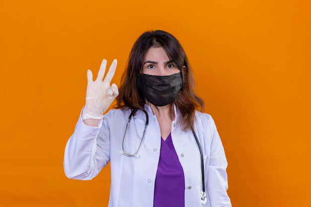 Vrouw arts van middelbare leeftijd dragen witte jas in zwarte beschermende gezichtsmasker en met stethoscoop camera kijken met vertrouwen expressie doen ok teken staande over oranje achtergrond