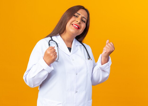 Vrouw arts van middelbare leeftijd in witte jas met stethoscoop blij en opgewonden balde vuisten staande over oranje muur