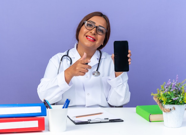 Vrouw arts van middelbare leeftijd in witte jas met stethoscoop bril met smartphone wijzend met wijsvinger naar voren glimlachend zittend aan de tafel over blauwe muur