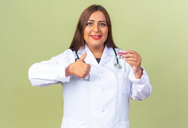 Vrouw arts van middelbare leeftijd in witte jas met stethoscoop die blaar vasthoudt met pillen die duimen laten zien glimlachend zelfverzekerd over groene muur