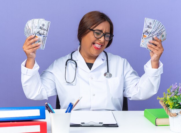 Vrouw arts van middelbare leeftijd in witte jas met stethoscoop met contant geld blij en opgewonden blij met haar succes zittend aan de tafel over blauwe achtergrond
