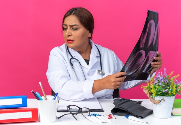 Vrouw arts van middelbare leeftijd in witte jas met stethoscoop met röntgenfoto opzij kijkend verward en erg angstig zittend aan de tafel met kantoormappen op roze