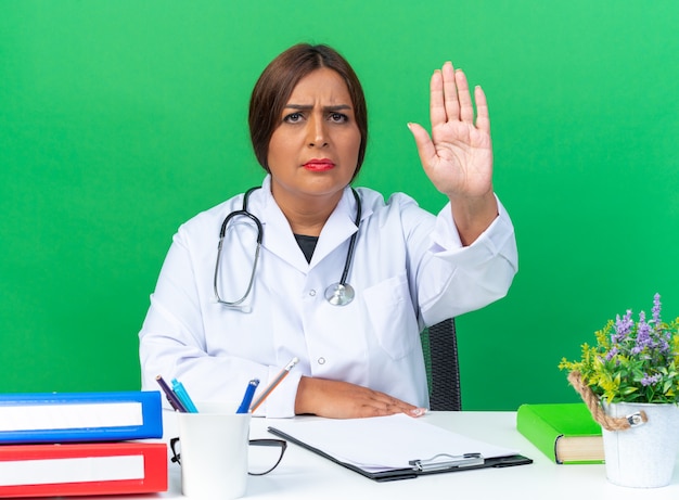 Vrouw arts van middelbare leeftijd in witte jas met stethoscoop met serieus gezicht maken stopgebaar met hand zittend aan tafel over groene muur