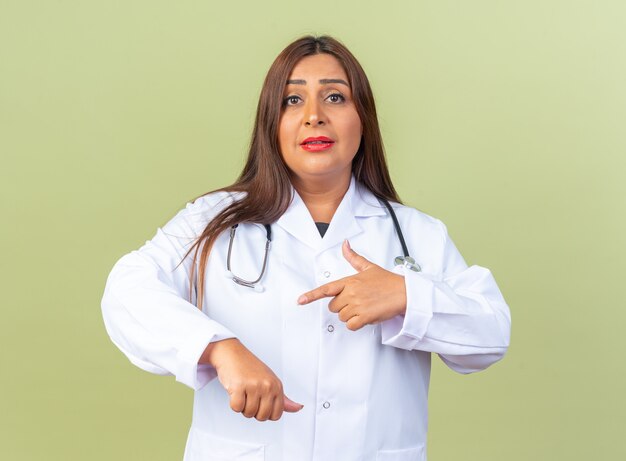 Vrouw arts van middelbare leeftijd in witte jas met stethoscoop wijzend met wijsvinger naar haar hand die herinnert aan de tijd die er zelfverzekerd uitziet op groen
