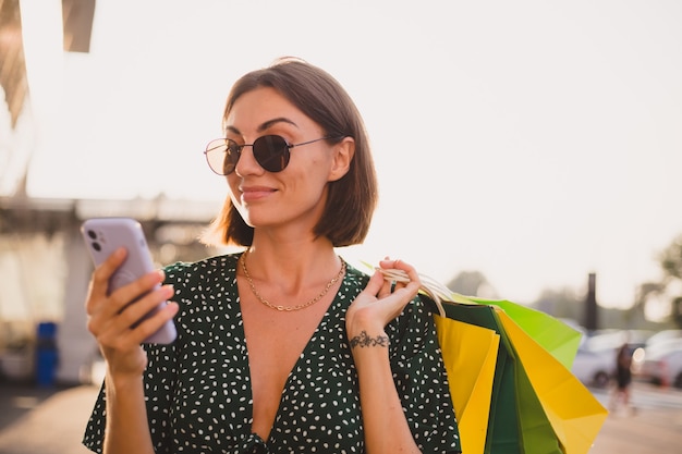 Vrouw bij zonsondergang met kleurrijke boodschappentassen en parkeerplaats bij winkelcentrum blij met mobiele telefoon