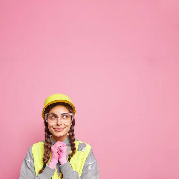 Vrouw bouwer assistent houdt handen bij elkaar kijkt naar boven peinzend denkt over de bouw van een nieuw gebouw draagt beschermende helm veiligheidsbril en uniform geïsoleerd over roze muur.