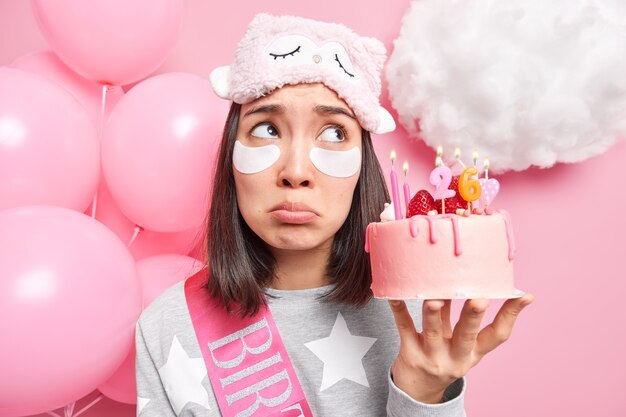 vrouw boven geconcentreerd met verstoorde uitdrukking past patches onder de ogen toe voelt zich eenzaam op verjaardag houdt smakelijke cake gekleed in zachte pyjama