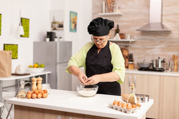 Vrouw breekt ei boven meel en maakt deeg voor bakkerijproducten. Oudere banketbakker die ei op glazen kom kraakt voor cakerecept in keuken, met de hand mengen, ingrediënten kneden en zelfgemaakte cake voorbereiden