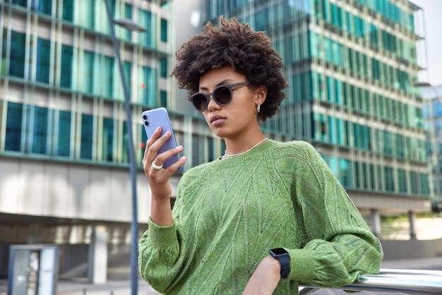 vrouw chat met vrienden in sociale netwerken surft op internetwebsite om interessante plekken in de stad te zoeken houdt mobiel draagt zonnebril en casual jumper poses in de buurt van kantoorgebouw