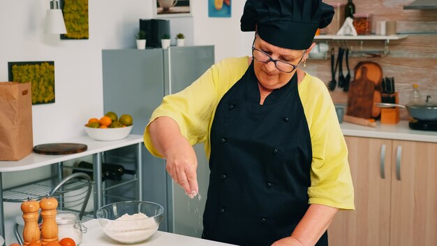 Vrouw chef-kok tarwebloem uit glazen kom nemen en zeven op tafel. Gepensioneerde senior bakker met bonete en uniform besprenkelen, zeven, verspreiden van nieuwe ingrediënten die zelfgemaakte pizza en brood bakken.