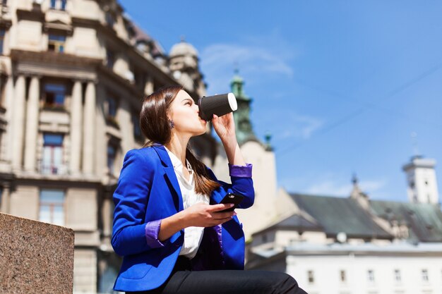 Vrouw controleert haar telefoon zitten met een kopje koffie op de straat