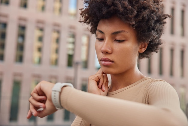 Gratis foto vrouw controleert pols na joggen training raakt nek aan kijkt op horloge gekleed in sportkleding staat buiten controleert haar gezondheid