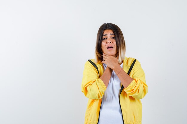 Vrouw die aan keelpijn in t-shirt, jas lijdt en ziek, vooraanzicht kijkt.