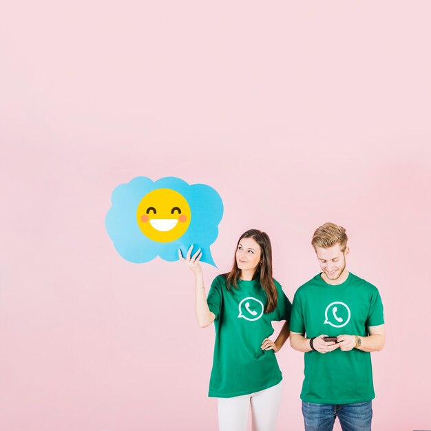 Vrouw die blauwe toespraakbel met lachende emoji houdt dichtbij de mens die cellphone gebruikt