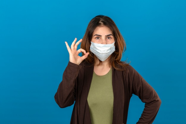 Vrouw die bruin vest in medisch beschermend masker draagt dat zeker kijkt die doend ok teken die zich over geïsoleerde blauwe muur bevinden