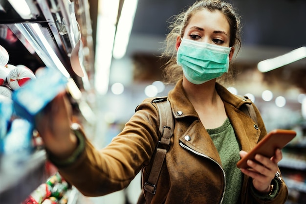 Vrouw die een beschermend masker draagt tijdens het gebruik van een mobiele telefoon en het kopen van voedsel in de supermarkt tijdens een virusepidemie