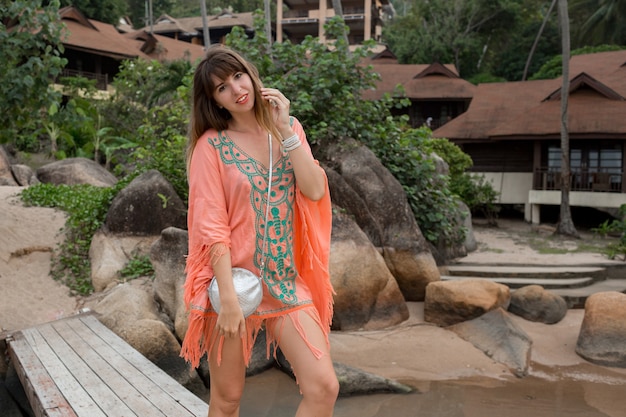 vrouw die een Boho-jurk draagt die op het strand loopt. Rotsen en palmbomen op achtergrond. Zomer mode.